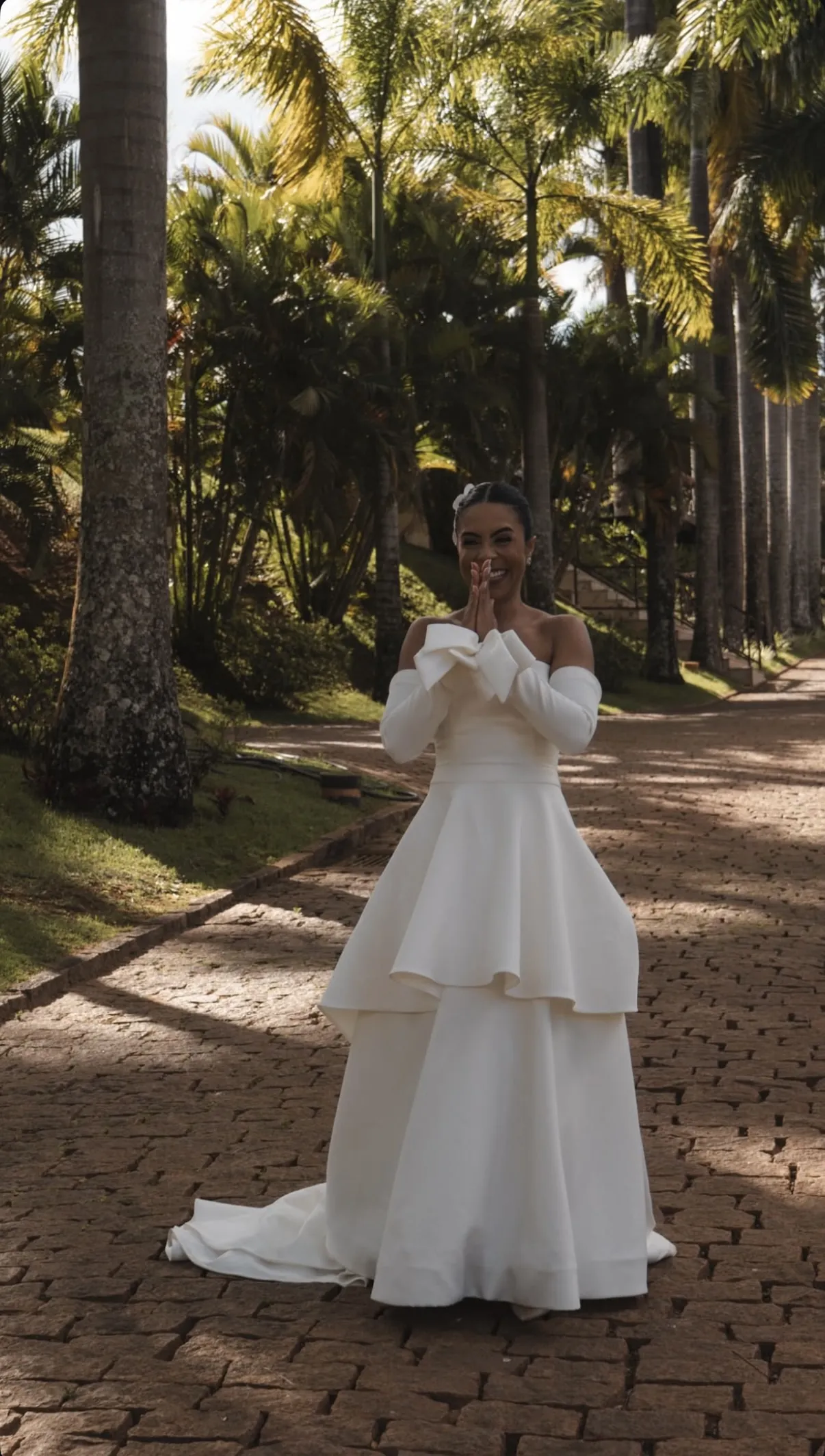 Noiva em alameda de palmeiras, com vestido de babados e luvas longas